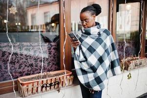 mujer africana con capa a cuadros y anteojos posó al aire libre y miró el teléfono móvil. foto