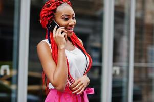 chica afroamericana de moda con pantalones rosas y rastas rojas posadas al aire libre con teléfono móvil. foto