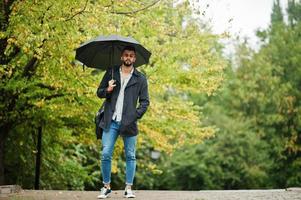 hombre de barba árabe alto de moda vestido con abrigo negro con paraguas y estuche posado en el día de la lluvia. foto