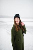 Funny girl wear on long green sweatshirt, jeans and black headwear, at frozen lake in winter day. photo