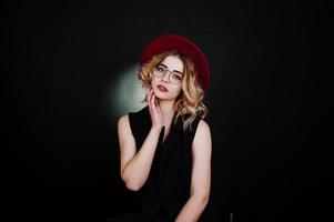 Studio portrait of blonde girl in black wear, red hat and glasses against dark background. photo