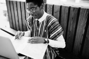 hombre africano con ropa tradicional y anteojos sentado detrás de una laptop en un café al aire libre y mirando el mapa de áfrica y ghana en su cuaderno. foto