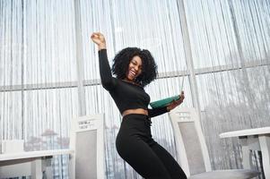 Happy and exults winner fashionable young beautiful african american business woman with afro hairstyle wear in elegant black, stand with laptop at hands.