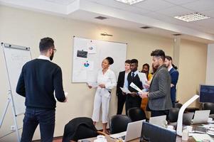 Mixed race business coach presenting report standing near whiteboard pointing on sales statistic shown on diagram and chart teach diverse company members gathered together in conference room. photo