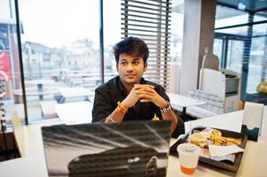 hombre indio con estilo sentado en un café de comida rápida contra su computadora portátil. foto