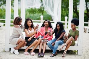 Group of five african american girls relaxing at beautiful poolside cabana beside luxury resort and looking at mobile phones. photo