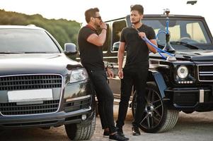 Two asian brothers man wear on all black posed near suv cars and smoke hookah. photo