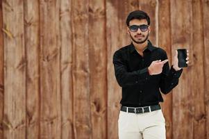 un joven indio informal con pantalones negro y gafas de sol posado sobre un fondo de madera que se muestra en el teléfono móvil. foto