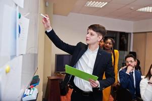 Mixed race business coach presenting report standing near whiteboard pointing on sales statistic shown on diagram and chart teach diverse company members gathered together in conference room. photo