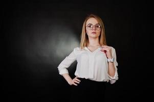 Studio portrait of blonde businesswoman in glasses, white blouse and black skirt against dark background. Successful woman and stylish girl concept. photo