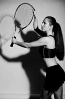 Black and white portrait of beautiful young woman player in sports clothes holding tennis racket while standing against white background. photo