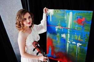 Pretty young gilrl musician in white dress with double bass next to her art painting. photo