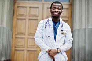 African american doctor male at lab coat with stethoscope outdoor against clinic door. photo