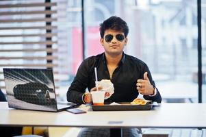 hombre indio elegante con gafas de sol sentado en un café de comida rápida contra su computadora portátil y muestra el pulgar hacia arriba. foto