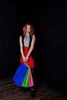 Portrait of an attractive redheaded girl in sleveless jacket, striped blouse and red trousers posing with multicolored packages next to the brick wall. Shopping spree. photo