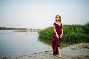 mujer rubia sensual descalza con vestido rojo marsala posando contra el lago con juncos. foto