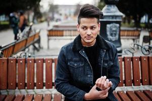 Handsome and fashionable indian man in black jeans jacket posed outdoor, sitting on bench. photo