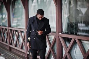 elegante caballero afroamericano con elegante chaqueta negra, sosteniendo un bastón retro como matraz de caña o bastón basculante con mango de bola de diamante dorado. rico hombre afro de moda. foto