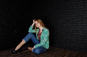 chica rubia con estilo en chaqueta y jeans contra la pared de ladrillo negro en el estudio. foto