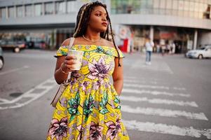 Cute small height african american girl with dreadlocks, wear at coloured yellow dress, walking at crosswalk against trade center with cup of coffee at hand. photo