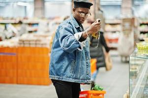 elegante hombre afroamericano casual con chaqueta de jeans y boina negra sosteniendo una canasta y mirando una botella de vino, comprando en el supermercado. foto