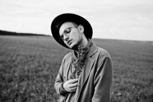 Stylish man in glasses, brown jacket and hat posed on green field. photo