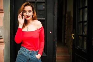 Attractive redhaired woman in eyeglasses, wear on red blouse and jeans skirt posing at street against old wooden door. photo