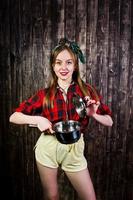 Young funny housewife in checkered shirt and yellow shorts pin up style with saucepan on wooden background. photo