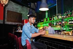 Portrait of handsome successful bearded south asian, young indian freelancer in blue jeans shirt sitting in night club against bar counter with cocktail and having a rest. photo