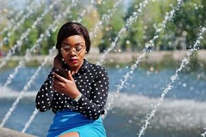 Stylish african american model girl in glasses, blue skirt and black blouse posed outdoor background fountains and looking on mobile phone. photo