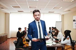 Face of handsome arabian business man, holding clipboard on the background of business peoples multiracial team meeting, sitting in office table. photo