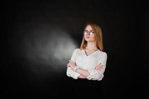 Studio portrait of blonde businesswoman in glasses, white blouse and black skirt against dark background. Successful woman and stylish girl concept. photo