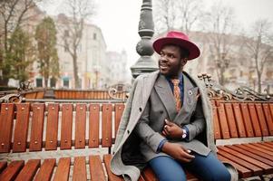 Stylish African American man model in gray coat, jacket tie and red hat posed at bench. photo