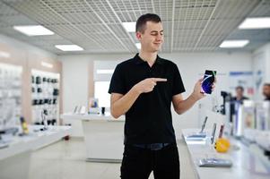 vendedor hombre consultor profesional de telefonía móvil en tienda de tecnología o tienda sostenga un nuevo teléfono inteligente a mano y muestre el dedo a la pantalla. foto