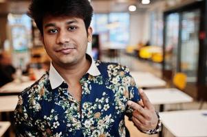 Close up portrait of young male indian freelancer in fast food cafe, handsome head of asian man wear in shirt at comfortable coffee shop shows two fingers sign. photo
