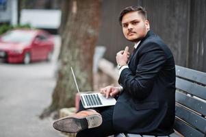 hombre de negocios indio con estilo en ropa formal sentado en un banco con una computadora portátil. foto