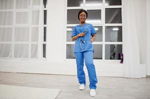 retrato de una joven y feliz doctora afroamericana pediatra con uniforme azul y estetoscopio contra la ventana del hospital. cuidado de la salud, médico, especialista en medicina - concepto. foto