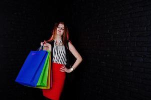 Portrait of an attractive redheaded girl in sleveless jacket, striped blouse and red trousers posing with multicolored packages next to the brick wall. Shopping spree. photo