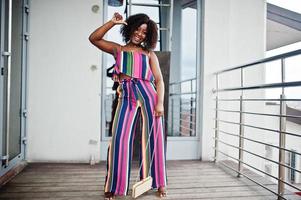 Fashionable african american woman in pink striped jumpsuit, with handbag posed. photo