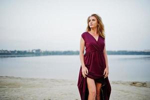 mujer rubia sensual descalza con vestido rojo marsala posando contra el lago en la arena. foto