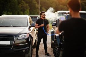 dos hermanos asiáticos se visten de negro y posan cerca de autos todoterreno y fuman narguile. foto