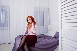 Gorgeous red haired girl in pink blouse and red skirt with glass of wine at hand on the bed at room. photo