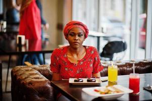 Stylish african woman in red shirt and hat posed indoor cafe, eat chocolate dessert cakes. photo