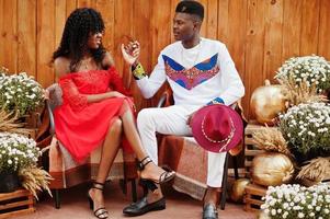 Stylish trendy afro france couple posed together at autumn day. Black african models in love sitting against wooden decoration with flowers and pumpkins. photo