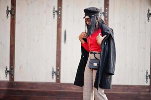 chica de moda afroamericana con abrigo y gorra de repartidor de periódicos posada en la calle. foto