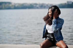 Modelo delgado afroamericano de piel oscura posado en pantalones cortos negros y chaqueta de jeans contra el mar. foto