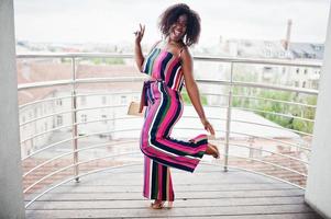 mujer afroamericana de moda y feliz con mono rosa a rayas, con bolso de mano posado en el balcón francés. foto