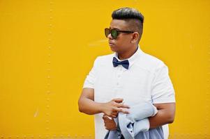 Close up portrait of stylish arabian man at sunglasses and bow tie posed against yellow background with mobile phone. Arab model boy. photo