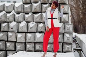 mujer afroamericana con pantalones rojos y chaqueta de abrigo de piel blanca posada en el día de invierno contra el fondo de piedra nevada. foto