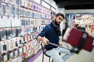 Indian man customer buyer at mobile phone store making selfie by smartphone on monopod stick. South asian peoples and technologies concept. Cellphone shop. photo